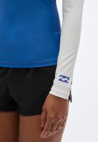 Μπλε μακρυμάνικη μπλούζα για προστασία από τον ήλιο με λευκά μανίκια, που έχει ένα μπλε λογότυπο κύματος στη μανσέτα. Μαύρο σορτς είναι ορατό στο κάτω μέρος. Λείο ύφασμα.