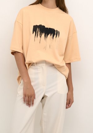T-shirt oversize couleur pêche en coton doux, avec un motif en coup de pinceau noir au centre. Associé à un pantalon de couleur claire.