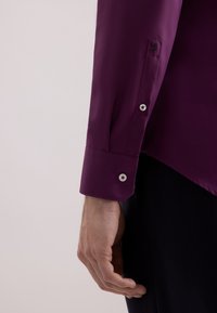 La mano sinistra di un uomo accanto al polsino di una camicia a maniche lunghe viola scuro con bottoni bianchi, con un piccolo dettaglio ricamato di rosa nero sulla manica.
