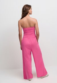 Stropløs jumpsuit i knaldrosa, med en tætsiddende overdel og brede, flydende bukseben. Materialet virker blødt med en glat tekstur.