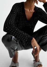 Suéter negro de punto con escote en V, adornado con embellishments cuadrados plateados. Combinado con unos pantalones de vinilo negro brillantes y tacones strappy con joyas.