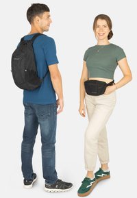 Schwarzer Rucksack mit Netz-Akzenten auf dem Rücken des Mannes; Frau mit grünem Bauchtasche und hellbeigen Hosen. Beide tragen lässige Sneakers.