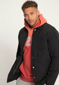 Giacca nera trapuntata con bottoni a pressione sopra un hoodie rosso. Presenta una tasca sul petto e un tessuto testurizzato. Pantaloni grigi visibili sotto.
