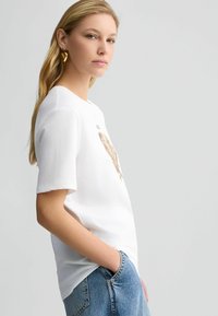 T-shirt in cotone bianco con scollo rotondo e maniche corte, con un design grafico dorato sul davanti. Abbinato a denim blu chiaro.