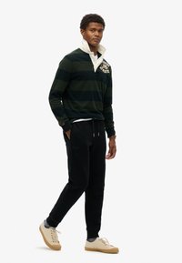 Donkergroene en zwarte gestreepte rugbyshirt met een witte kraag, gecombineerd met zwarte joggingbroek en tan schoenen, met een gestructureerde stof.