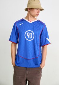 Zils Nike krekls ar šaha rakstu, sarkaniem akcentiem un apaļu "90" logo. Iezīmēts ar baltu apkakli un īsām piedurknēm.