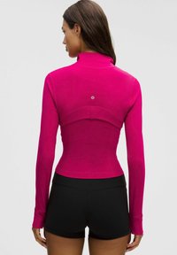 Femeie îmbrăcată într-un top magenta ajustat cu mâneci lungi și orificii pentru degete, purtând pantaloni scurți negri, văzută din spate pe un fundal simplu.