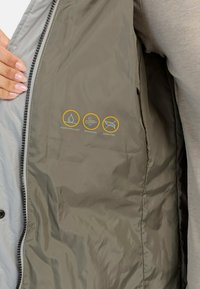 Vandafvisende, vindbestandig jakke uden dun i dæmpet olivengrøn med en glat tekstur og indvendige pleje-symboler trykt i grå og gul.
