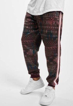 Unterkörper in weinroten gemusterten Jogginghosen mit seitlichen Streifen und weißen Sneakers, stehend auf einem einfarbigen Hintergrund.