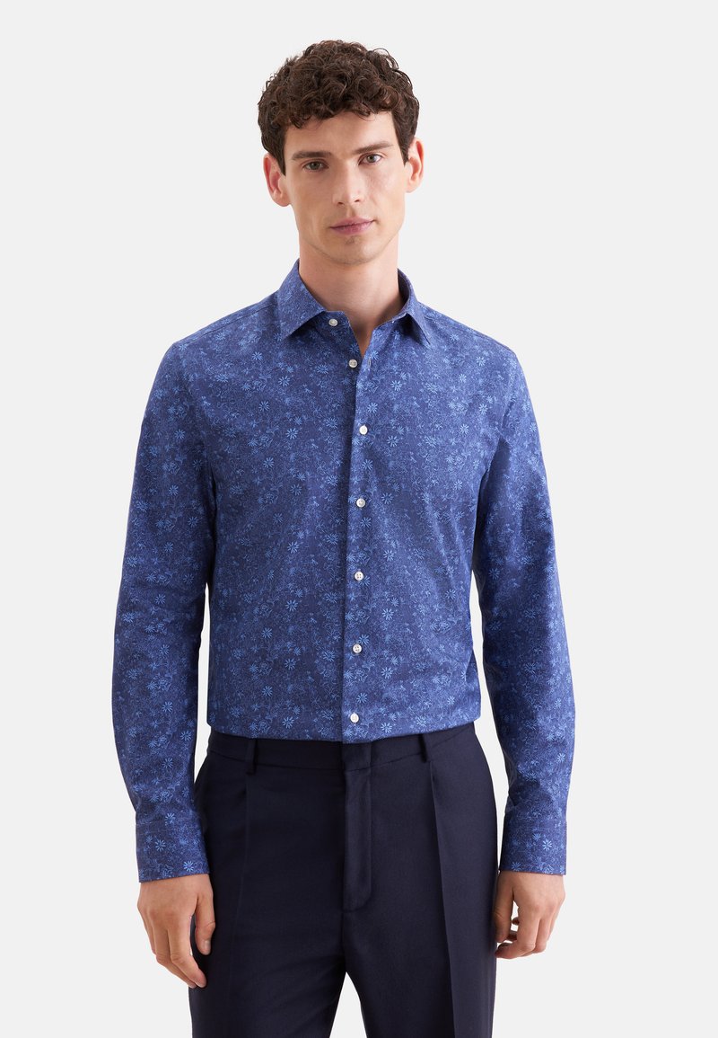 Homme aux cheveux bouclés portant une chemise bleue fleurie ajustée à boutons et un pantalon bleu marine foncé, debout devant un fond uni.