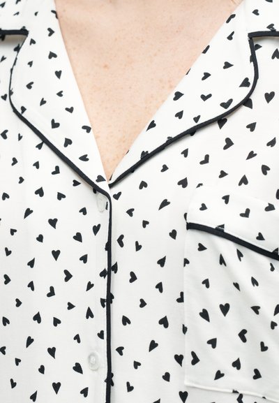 Λευκό πάνω μέρος πυτζάμας με μαύρα μοτίβα καρδιάς, μαύρη επένδυση κατά μήκος του γιακά, της πλακέτας με κουμπιά και της τσέπης στο στήθος, που φοριέται από άτομο.