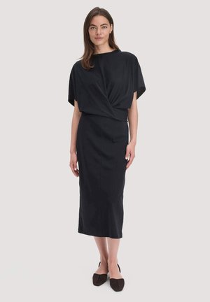 Vrouw staat, gekleed in een zwarte midi-jurk met een gedrapeerde top en korte mouwen, gecombineerd met zwarte puntige ballerina's op een effen achtergrond.