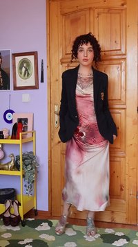 Person with curly hair wearing a pink and cream embellished dress, black blazer, and silver heels stands in front of a wooden door indoors.