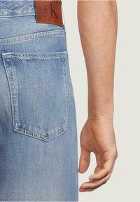 Jeans in denim azzurro chiaro con una tasca posteriore di forma rettangolare, una toppa in pelle marrone sulla cintura e una mano visibile di lato.