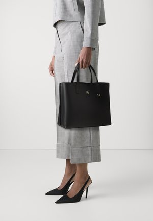 Schwarze Leder-Tote mit zwei Griffen, auf der Vorderseite mit einem goldenen Logo versehen. Kombiniert mit einem grauen karierten Outfit und schwarzen High Heels.