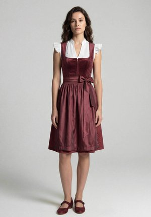 Frau steht und trägt ein knielanges bordeauxrotes Kleid mit einer Schleife an der Taille, eine weiße, kurzärmelige Bluse mit Rüschen und passende Schuhe.