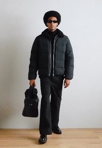 Sort pufferjakke med lynlås foran og krave, sort turtleneck, sorte flagrende bukser og sorte sko, mens der holdes en sort taske.