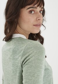 Grøn strikket sweater med en glat hvid krave, tætsiddende pasform og lange ærmer. Har et diskret mønster og blødt stof.