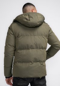 Jachetă puffer verde măsliniu cu un glugă mare, design matlasat și manșete elastice. Prezintă o textură netedă și un branding subtil pe mânecă.