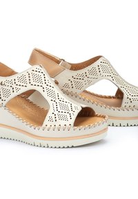 Sandali in pelle perforata beige con punta aperta, cinturino alla caviglia e cuciture testurizzate su una suola in gomma bianca.