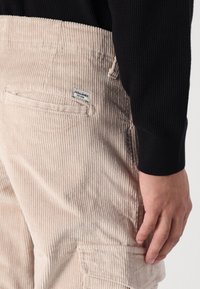 Beige fløjlsbukser med ribbet tekstur, der har en baglomme og et lille mærke. Båret med en sort tekstureret langærmet top.
