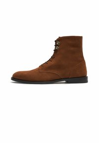 Botas de tobillo de ante marrón con punta redondeada, seis ojales de metal para los cordones y una suela de goma plana. Textura suave y diseño minimalista.