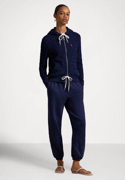 Ženska, oblečena v temno modro kapuco s patentnim trakom, pleteno z vzorcem vrvice in belimi vrvicami za zategovanje, ujemajoče se športne hlače in bele sandale, stoji z rokami v žepih.