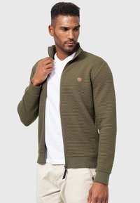 Chaqueta de cremallera verde olivo con superficie texturizada, puños acanalados y un pequeño parche de logo marrón en el pecho. Llevada sobre una camiseta blanca.