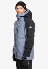 Blaue und schwarze Skijacke mit Kapuze, ausgestattet mit einem Reißverschluss vorne, verstellbaren Manschetten und weichem Material. Logo auf dem linken Ärmel zu sehen. Fäustlinge sichtbar.