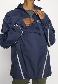 Mörkblå vindjacka med halvdragkedja, elastiska ärmar, reflexränder och justerbar nederkant. Tillverkad av lätt, vattenavvisande material.