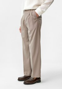 Beige plooibroek van lichtgewicht stof, met riemlussen en zijzakken, gecombineerd met bruine leren loafers.