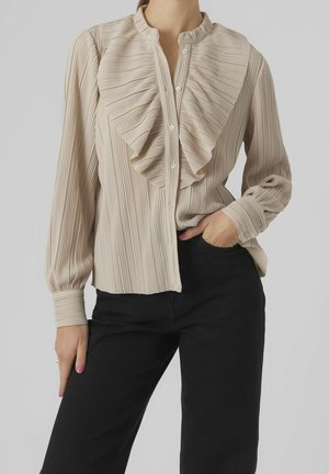 Beige geplooide blouse met lange mouwen en geruimde voorkant, gedragen ingestopt in een zwarte broek met hoge taille, één hand in de zak.