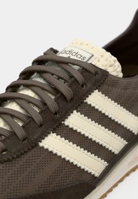 Brązowy zamszowy trampek z beżowymi skórzanymi paskami, teksturowaną tkaniną oraz plecionym kremowym językiem. Wyposażony w brązowe sznurowadła i gumową podeszwę.