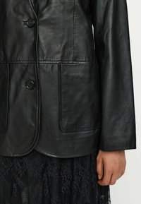 Zwarte leren blazer met een gladde textuur, voorzien van twee knopen, een ronde kraag en twee voorzakken. Gecombineerd met een zwarte kanten rok.