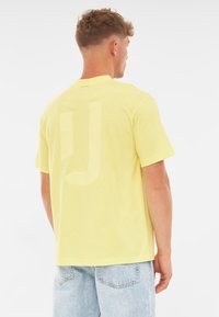 Żółta bawełniana koszulka z krótkim rękawem, z dużym, wyblakłym logo marki na plecach. Materiał wydaje się miękki i ma luźny krój.