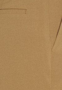 Tkanina v barvě béžové s hladkou texturou obsahuje malou šikmou kapsu a viditelné stehy podél švů.