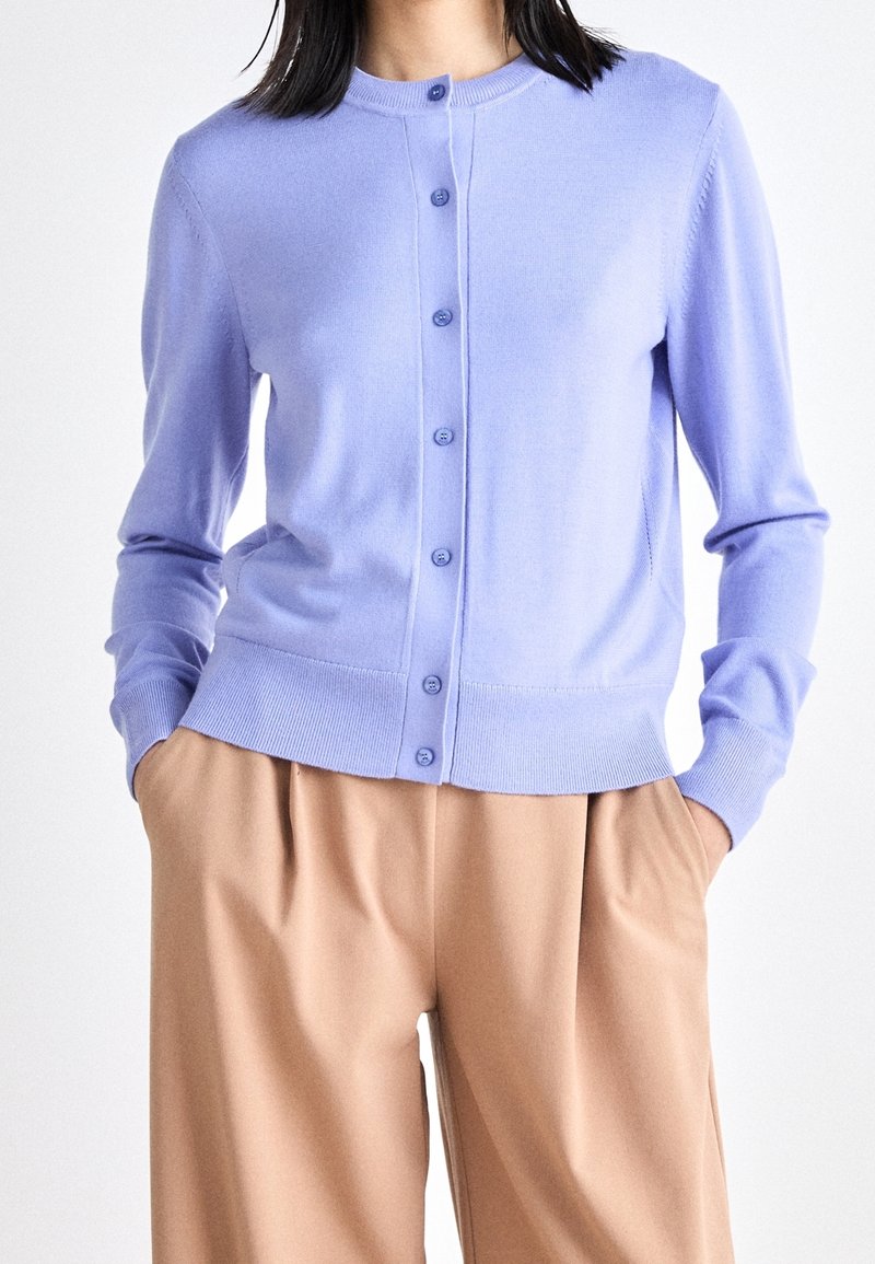 Femme portant un cardigan à boutons violet clair à manches longues et un pantalon plissé beige taille haute, les mains dans les poches.