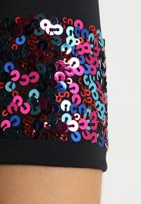 Černá látka se švem zdobeným vícebarevnými flitry (červené, růžové, modré a fialové), uspořádanými do vzoru, který vytváří strukturovaný detail.