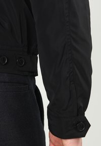 Sort letvægtsjakke med et tætsiddende design, elastisk talje og knapdetaljer på ærmerne. Glat tekstur med subtil glans.