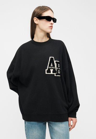 Nő fekete túlméretezett pulóverben, nagy fehér és fekete betűfoltokkal, világoskék farmerral, jobbra néz, fekete napszemüveget visel.