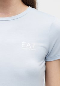 Κοντινό πλάνο ενός ατόμου που φοράει ανοιχτό μπλε κοντομάνικη μπλούζα με λευκό λογότυπο "EA7 Emporio Armani" στο στήθος.