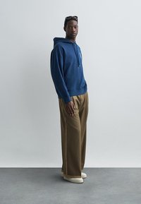 Sweat à capuche bleu marine avec poche avant, associé à un pantalon large beige avec des rayures sur les côtés. Le modèle porte des chaussures beiges et des lunettes de soleil.