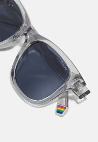 Ochelari de soare cu montură transparentă și lentile închise. Detaliu notabil: accente colorate în dungi de curcubeu pe braț. Formă pătrată distinctivă.