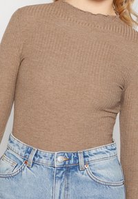 Maglietta lunga a maniche con righe marroni e scollatura ondulata, abbinata a jeans in denim azzurro chiaro a vita alta con bottoni e tasche.