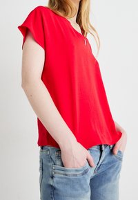 Punainen lyhythihainen paita sileästä kankaasta, V-aukolla ja väljällä istuvuudella, yhdistettynä vaaleansinisiin farkkuihin, joissa on etutaskut.