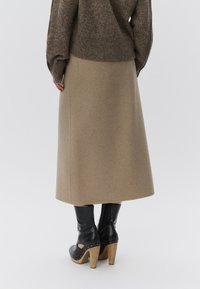 Μπεζ μάλλινη μίντι φούστα με ίσια σιλουέτα, σε συνδυασμό με μαύρες μποτάκια με τακούνι, που έχουν ξύλινη βάση και ασημένιες λεπτομέρειες.