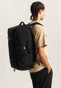 Zwarte rugzak met verstelbare banden, rechthoekige vorm en twee gespen aan de voorkant. Gemaakt van duurzaam materiaal en voorzien van een klein rood logodetail.