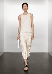 Helle beigefarbene, ärmellose Bluse kombiniert mit einem gestuften, knielangen Rock. Durchsichtige Stofftextur. Accessoire: weiße Häkel-Clutch. Schuhwerk: schwarze Sandalen.