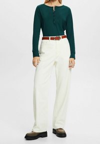 Langärmliges, dunkelgrünes Henley-Shirt mit Knopfdetails, kombiniert mit lockeren, weißen Hosen und einem braunen Gürtel. Klobige, braune Plateauschuhe sind ebenfalls zu sehen.