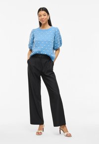 Suéter de punto azul cielo con puntos blancos texturizados, mangas abullonadas y pantalones negros de cintura alta. El modelo lleva sandalias de tacón ligero.